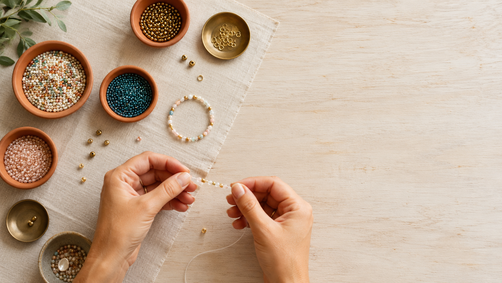 A pair of hands threading small glass beads onto a bracelet, materials spread across a linen tabletop